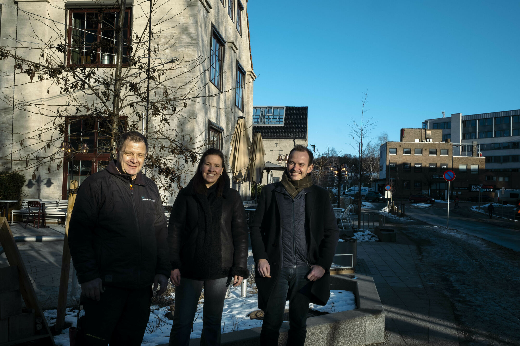 En kvinne og to menn oppstilt foran bygninger med solen i ansiktet